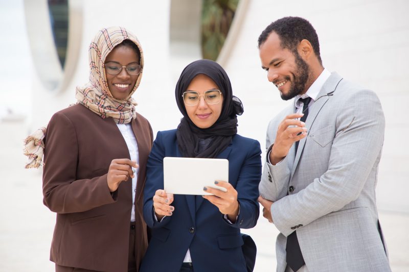 Front view of smiling managers using tablet outdoors. African American man and Muslim women looking at tablet. Technology concept